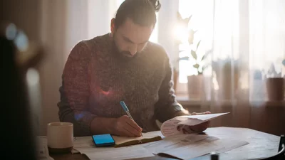 Mann mit Zopf schreibt Notizen in ein Notizbuch an einem Tisch mit Dokumenten und Smartphone