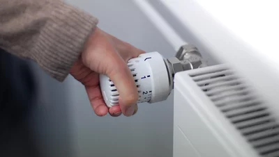 Hand dreht Thermostatventil an einem weißen Heizkörper zur Temperaturregelung