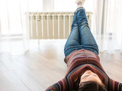 Frau liegt entspannt auf dem Boden mit den Füßen auf einem weißen Heizkörper vor einem Fenster