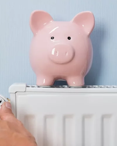 Gasheizung auf Wärmepumpe umrüsten Hand dreht Thermostat an weißem Heizkörper, daneben rosa Sparschwein auf hellblauem Hintergrund