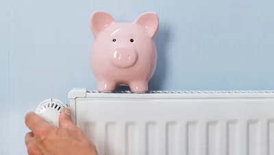 Gasheizung auf Wärmepumpe umrüsten – von Kosten bis Ablauf Hand dreht Thermostat an weißem Heizkörper, daneben rosa Sparschwein auf hellblauem Hintergrund