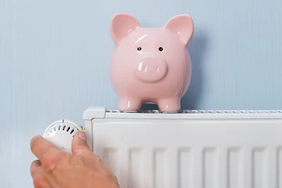 Hand dreht Thermostat an weißem Heizkörper, daneben rosa Sparschwein auf hellblauem Hintergrund