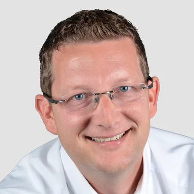 Porträt eines lächelnden Mannes mit Brille und weißem Hemd vor neutralem Hintergrund