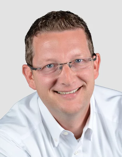 Porträt eines lächelnden Mannes mit Brille und weißem Hemd vor neutralem Hintergrund