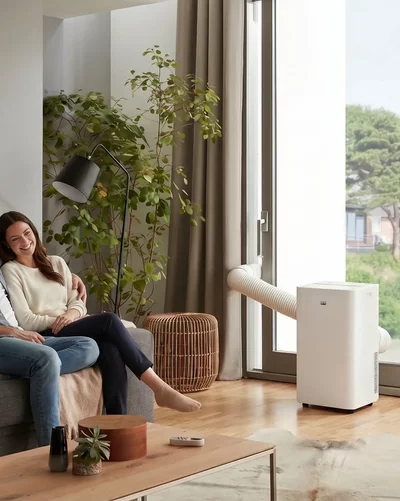 A couple relaxes on the sofa next to a white Remko air conditioner with exhaust hose near the window.